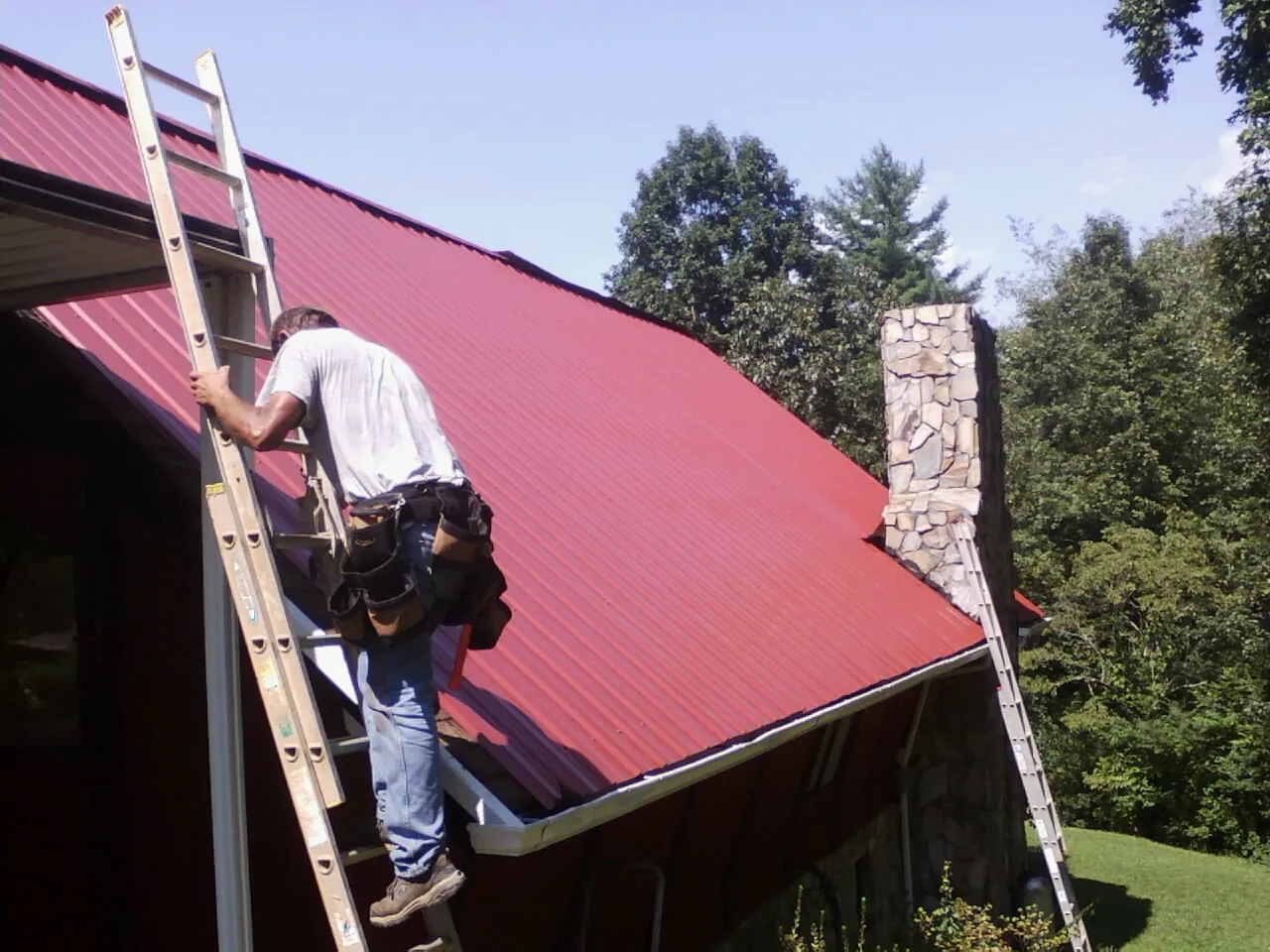 Roofer on a ladder beside a red metal roof and stone chimney — Soffit & Fascia Repair by Roofing Brothers in Westphalia